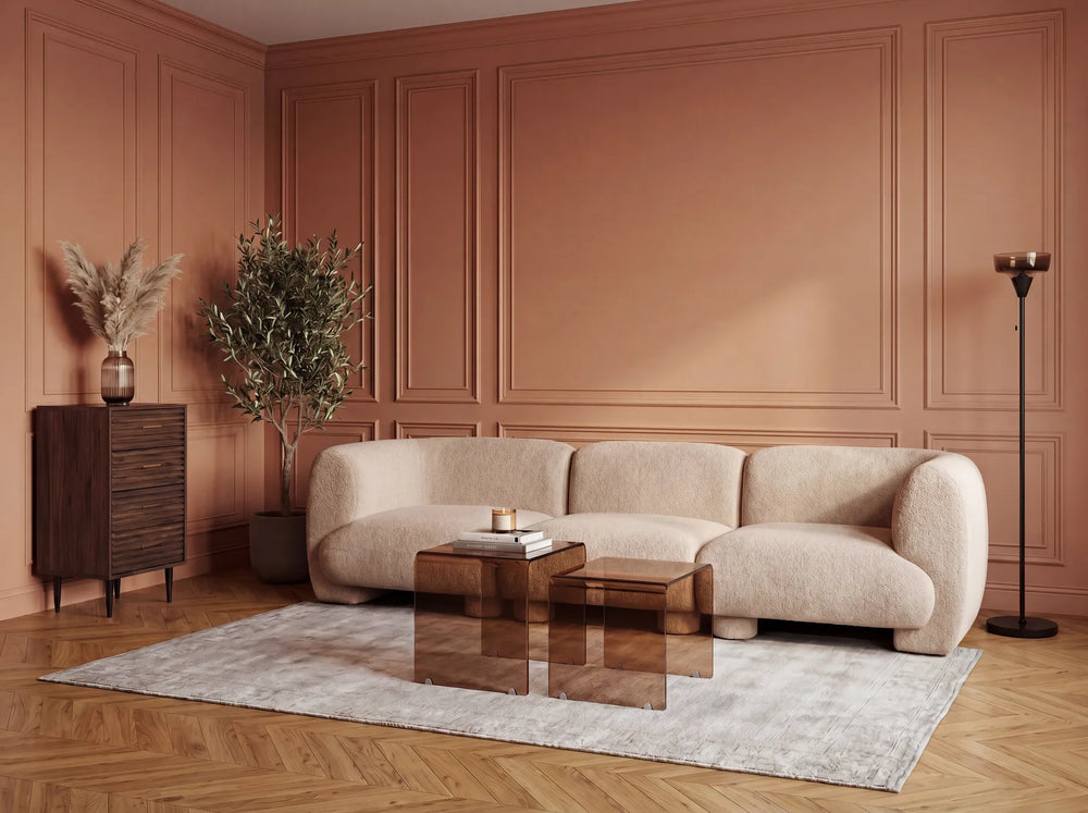 A modern living room scene featuring a curved, low-profile beige boucle sofa as the main product, styled on a light gray rug with two translucent amber glass coffee tables, a dark wood sideboard, and a tall floor lamp against a terracotta paneled wall.