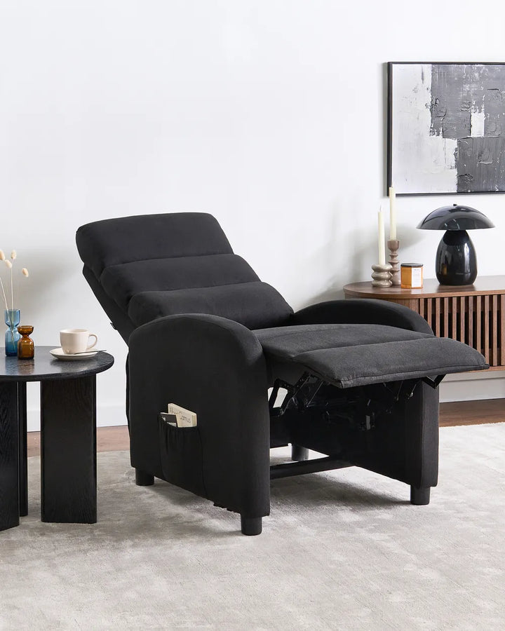 A black fabric recliner chair with the footrest extended, shown in a modern living room setting next to a small black side table and a wooden cabinet.