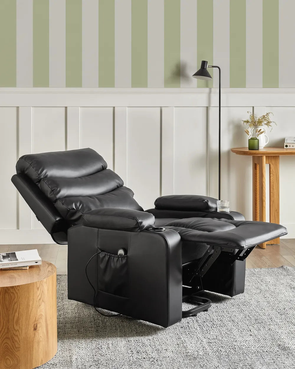 A black leather recliner chair with the footrest extended, shown in a styled room setting with striped wallpaper, a floor lamp, a side table, and a rug.
