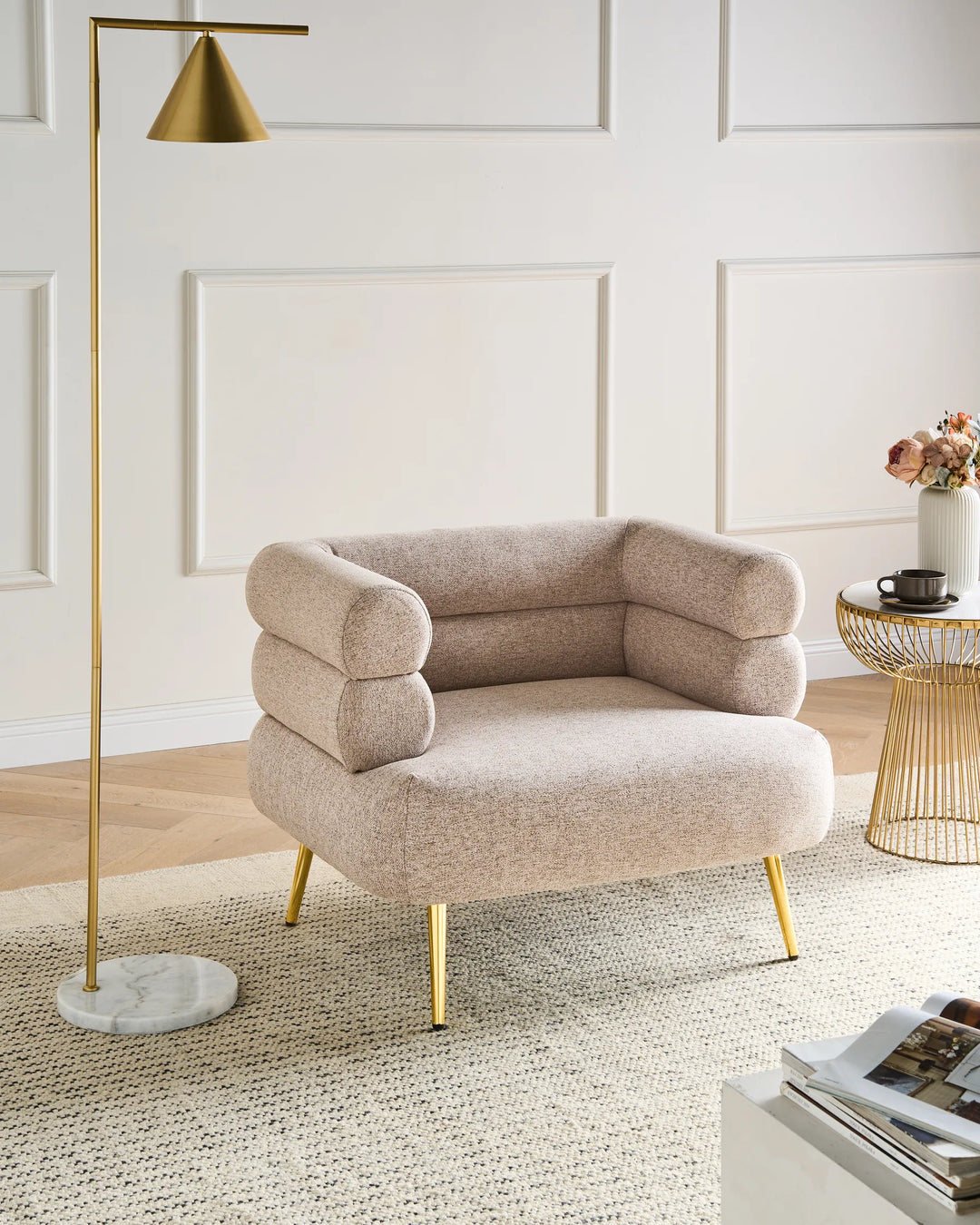 A modern upholstered accent chair in a warm beige boucle fabric with gold metal legs, styled in a bright room next to a gold floor lamp and a wire side table.