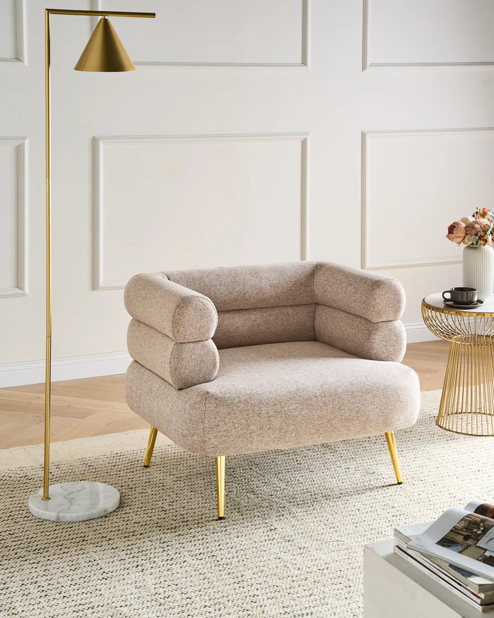 A modern upholstered accent chair in a warm beige boucle fabric with gold metal legs, styled in a bright room next to a gold floor lamp and a wire side table.