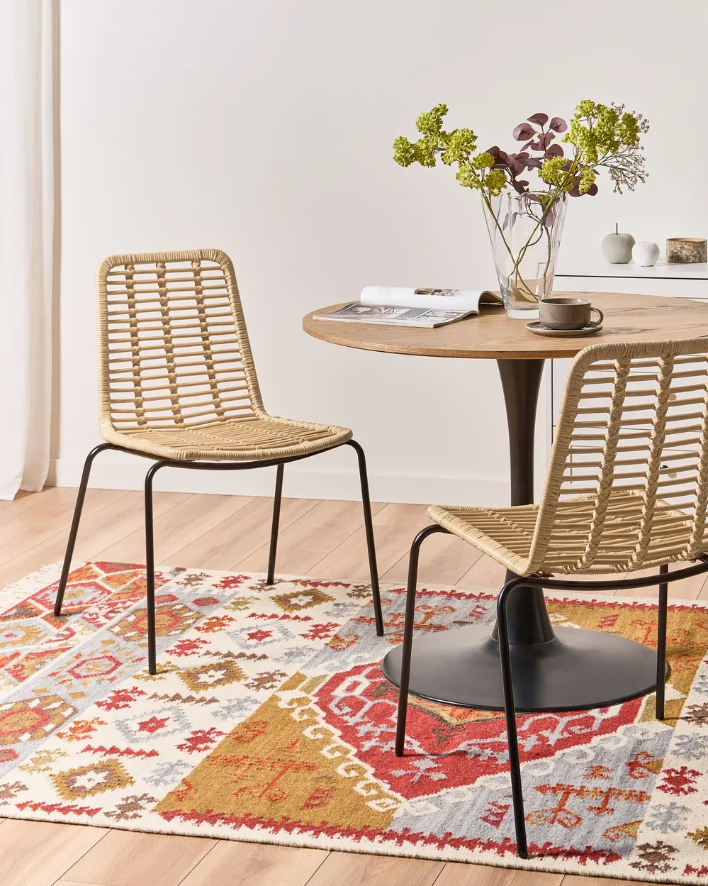 Two woven rattan dining chairs with black metal legs are shown in a bright dining room setting next to a round wooden table on a patterned rug.