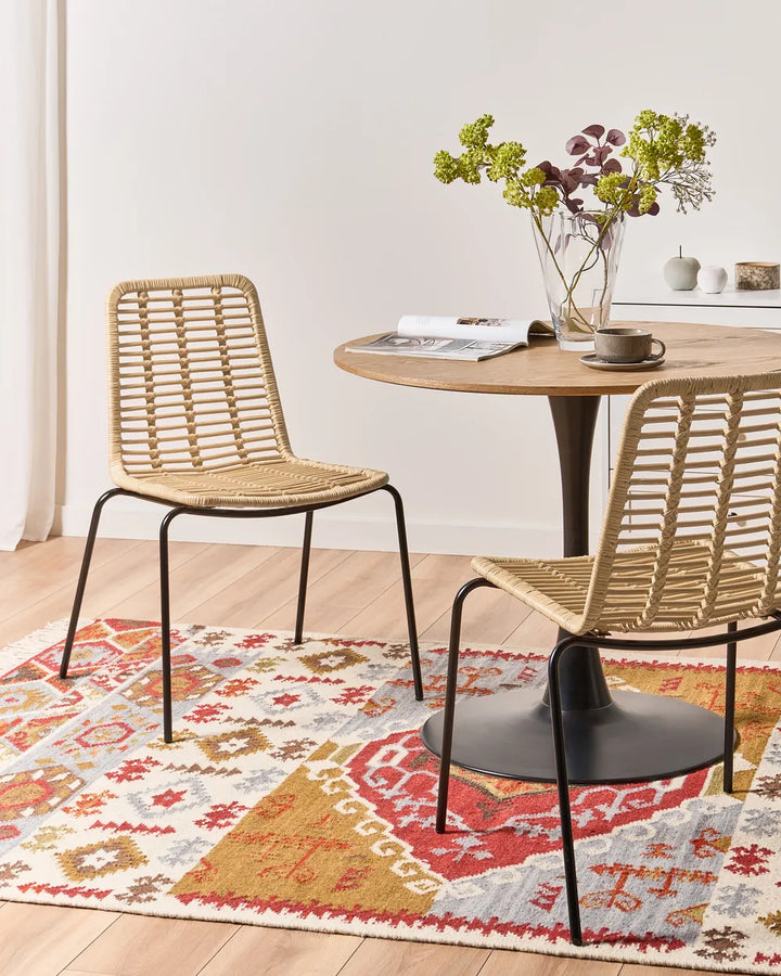 Two woven rattan dining chairs with black metal legs are shown in a bright dining room setting next to a round wooden table on a patterned rug.