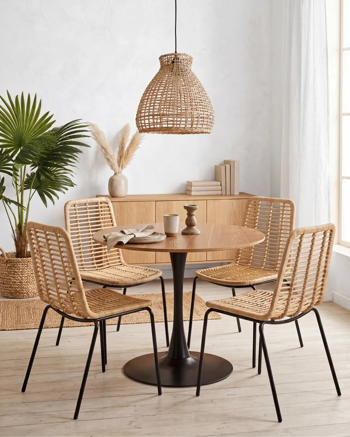 A round wooden dining table with a black pedestal base, surrounded by four woven rattan chairs with black metal legs, in a bright, modern dining room setting with a wicker pendant light and a wooden credenza.