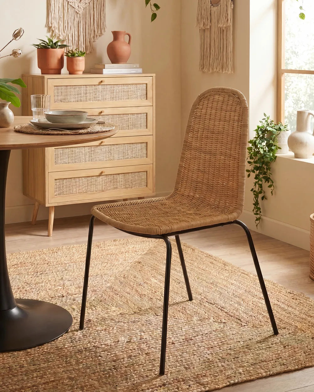 A woven rattan dining chair with a black metal frame, styled in a warm, bohemian dining room next to a wooden table and dresser.
