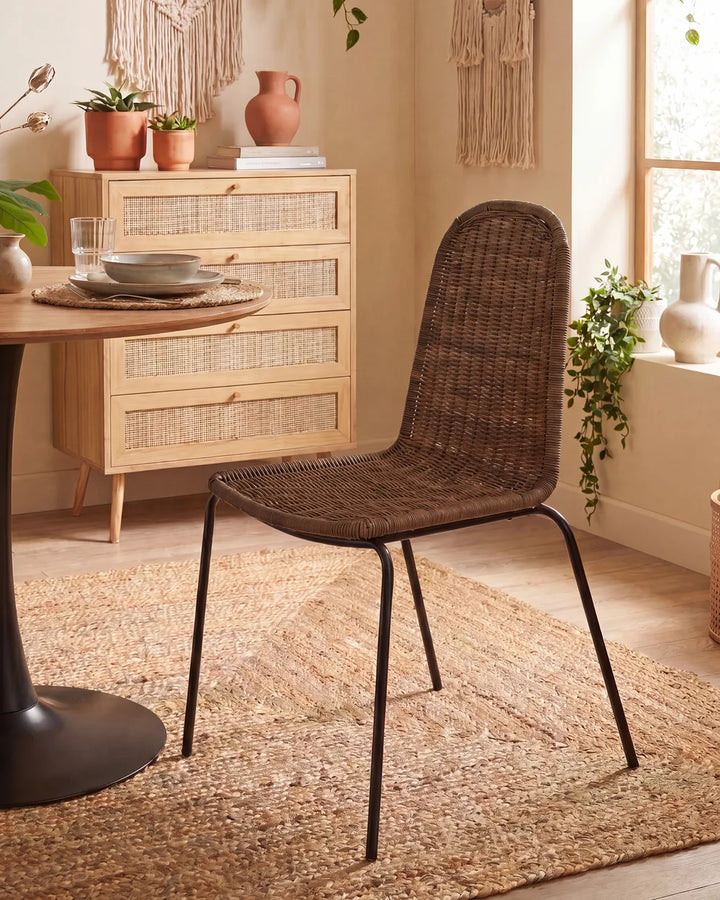 A dark brown woven rattan dining chair with black metal legs, positioned in a warm, boho-style dining room next to a wooden table and a light wood chest of drawers.