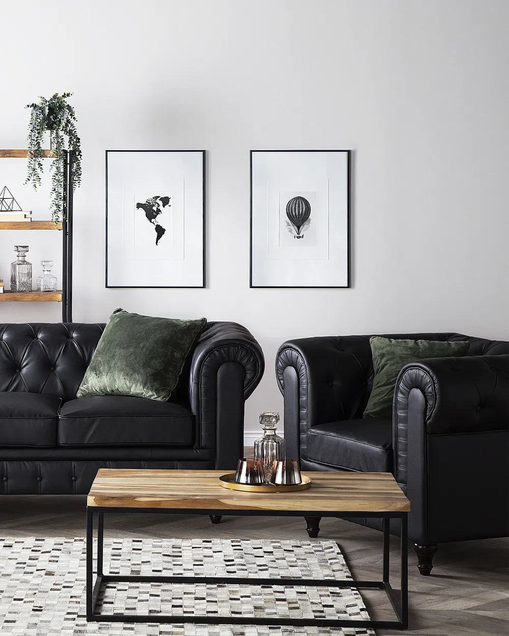 A styled living room featuring a black leather Chesterfield sofa and matching armchair with dark green velvet pillows, a wooden coffee table, and two framed black-and-white art prints on a white wall.
