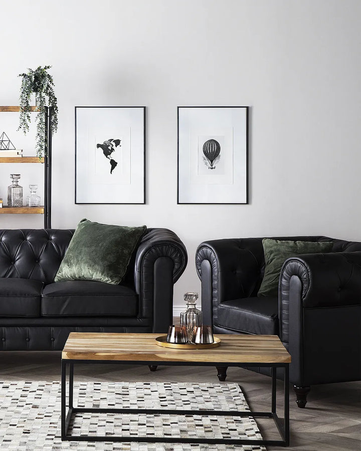 A styled living room featuring a black leather Chesterfield sofa and matching armchair with dark green velvet pillows, a wooden coffee table, and two framed black-and-white art prints on a white wall.