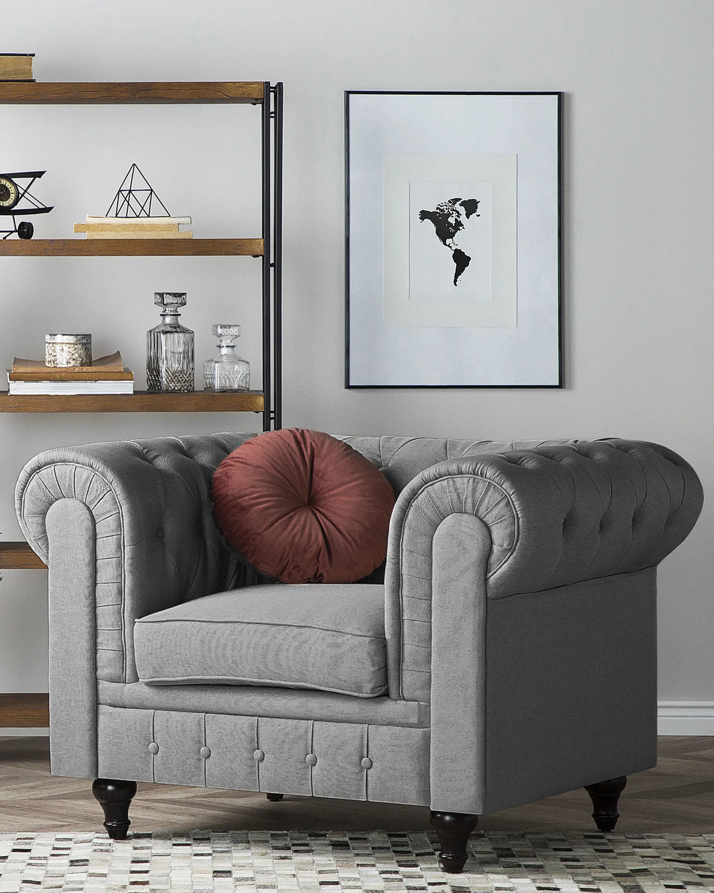 A gray tufted armchair with rolled arms and dark wooden legs, styled in a living room setting with a round rust-red throw pillow, a wooden bookshelf, and a framed world map print on the wall.