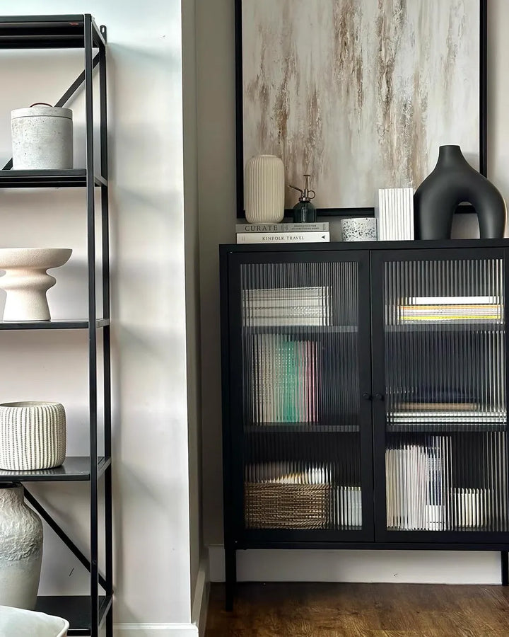 A modern black metal sideboard cabinet with ribbed glass doors, styled in a minimalist room next to a black shelving unit.