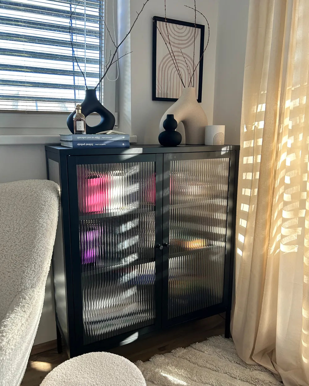 A modern black two-door cabinet with ribbed glass doors, styled in a sunlit room next to a window with blinds and a beige curtain.