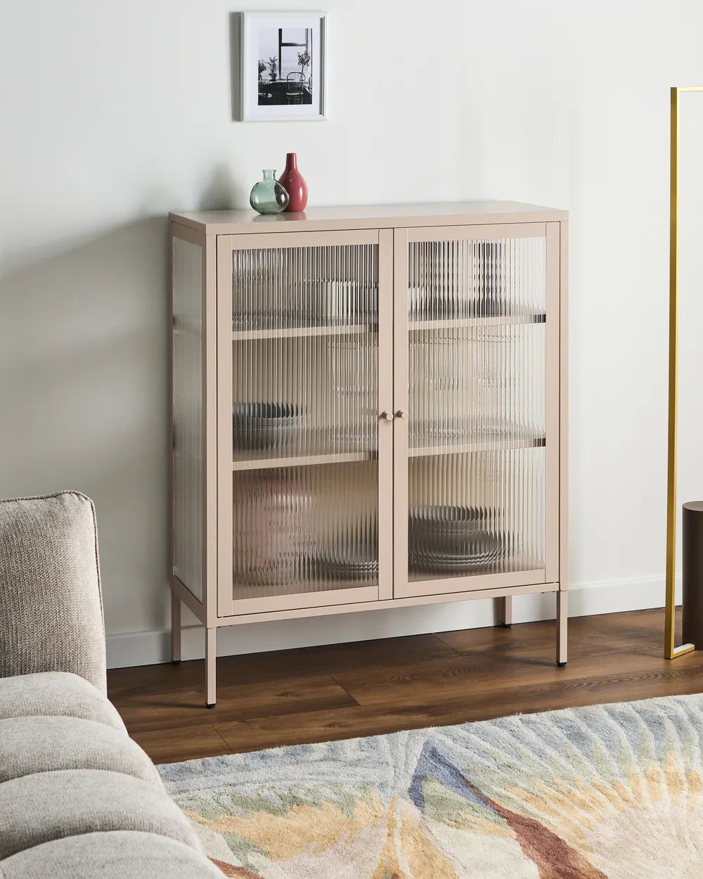 A light beige wooden sideboard cabinet with two ribbed glass doors, styled in a modern living room setting with a wooden floor, a gray sofa, and a patterned rug.