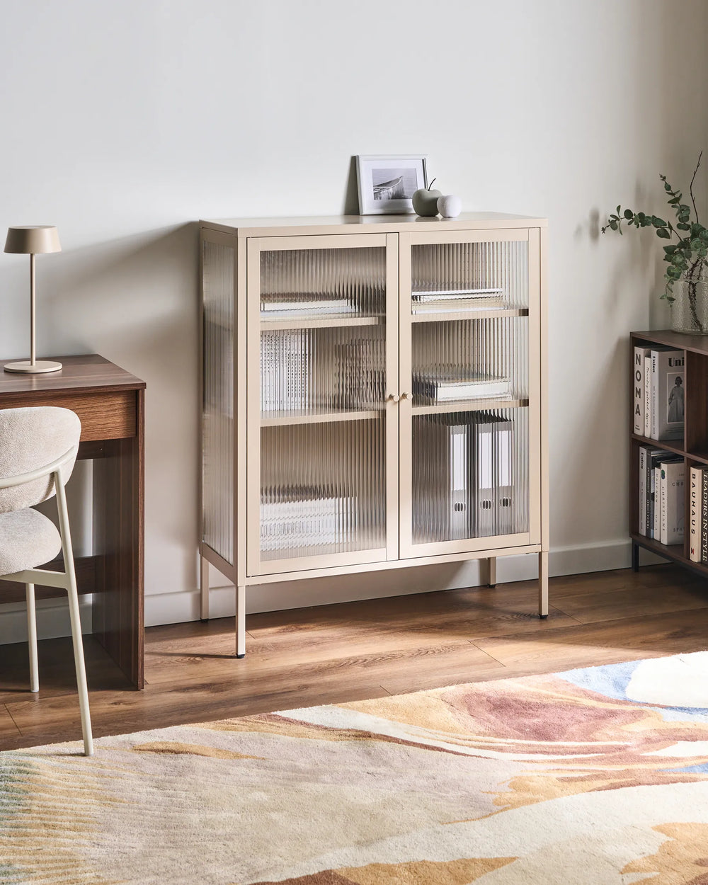 A tall, narrow side cabinet with two ribbed glass doors in a light beige wood finish, styled in a minimalist home office setting with a wooden desk, chair, and patterned rug.