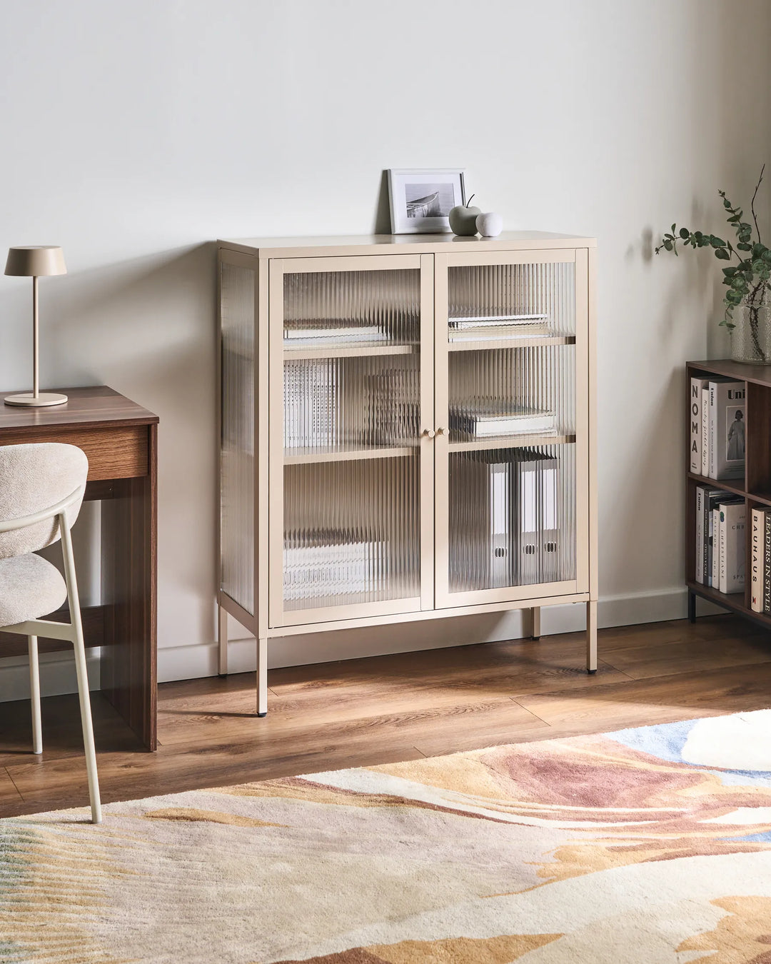 A tall, narrow side cabinet with two ribbed glass doors in a light beige wood finish, styled in a minimalist home office setting with a wooden desk, chair, and patterned rug.