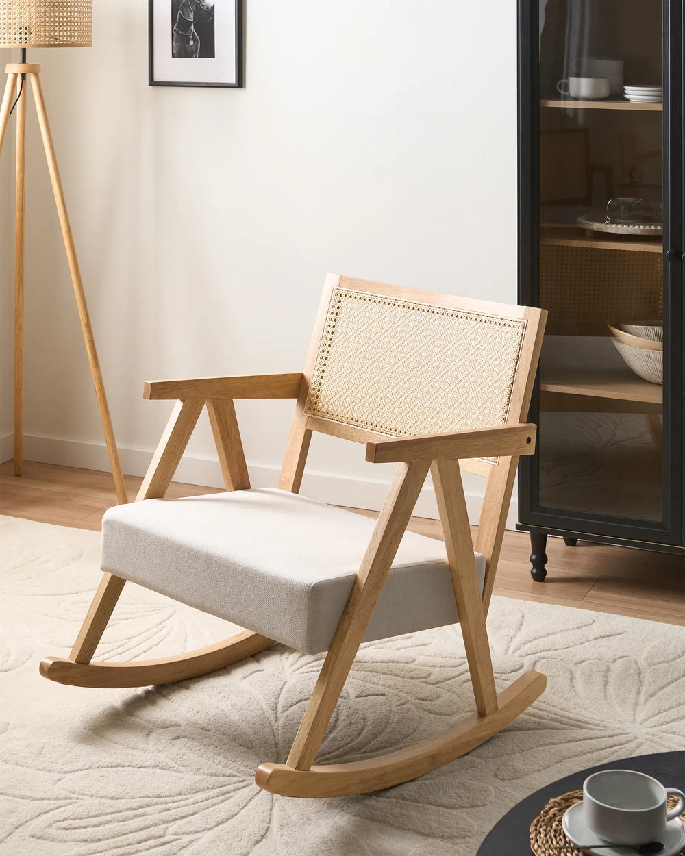 A modern wooden rocking chair with a woven cane backrest and a light beige upholstered seat, placed in a styled living room setting on a textured rug.
