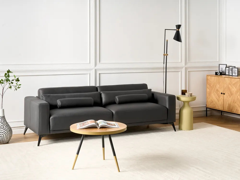 A modern black leather sofa in a styled living room setting with a round wooden coffee table, a gold side table, a black floor lamp, and a light wood cabinet against white paneled walls.