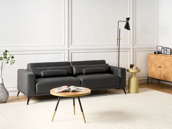 A modern black leather sofa in a styled living room setting with a round wooden coffee table, a gold side table, a black floor lamp, and a light wood cabinet against white paneled walls.
