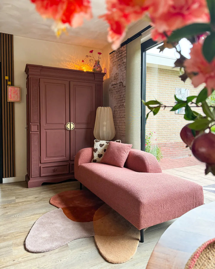 A dusty rose textured chaise lounge with matching pillows in a cozy, modern living room setting, featuring a large mauve wardrobe, abstract rugs, and a brick patio visible through a glass door.