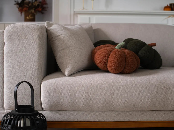 A light beige upholstered sofa in a styled interior, with a matching beige pillow and two fuzzy decorative pumpkins in rust orange and dark green resting on the seat.