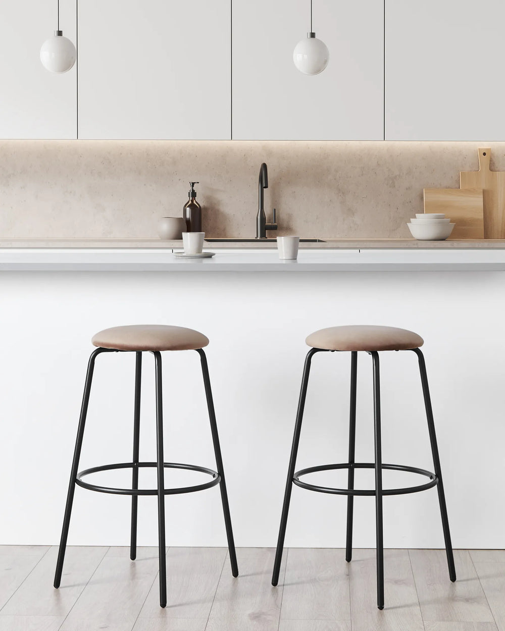 Two modern bar stools with round taupe-brown upholstered seats and black metal legs, positioned in front of a white kitchen island with a beige stone backsplash.