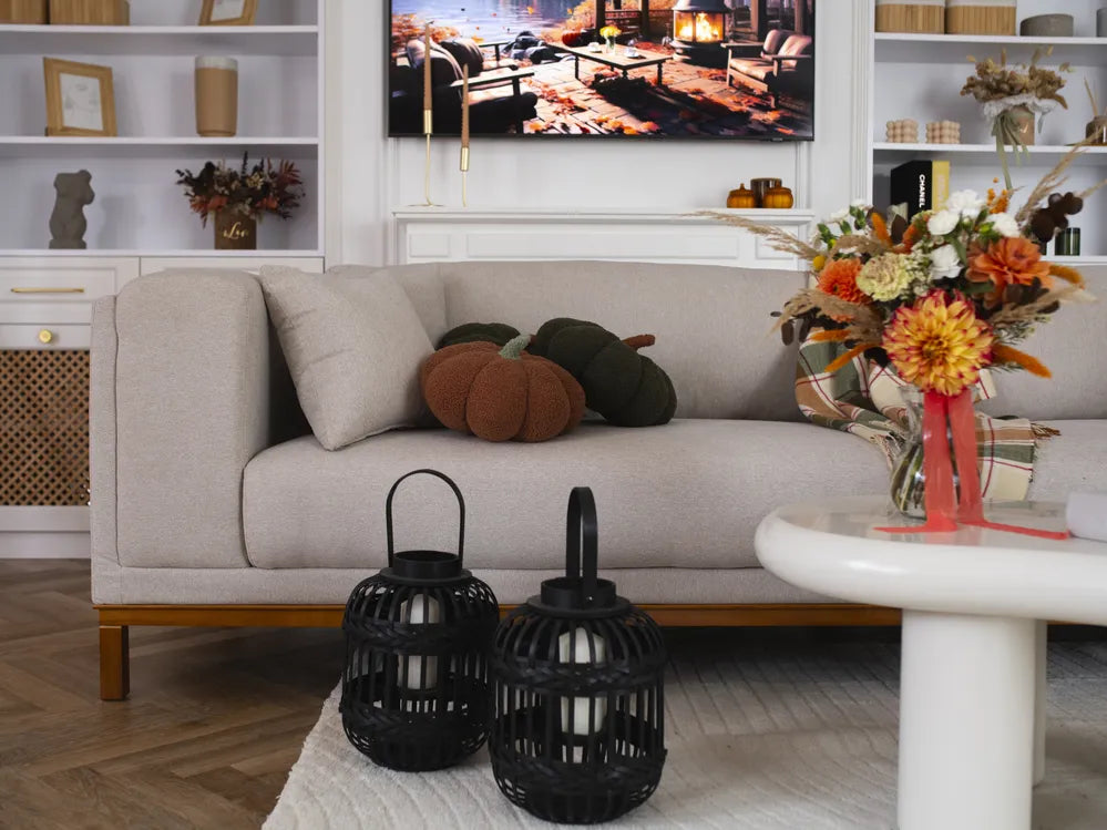 A styled living room scene featuring a light beige fabric sofa with decorative pumpkins, a white side table with a colorful floral arrangement, and two black lanterns in the foreground.