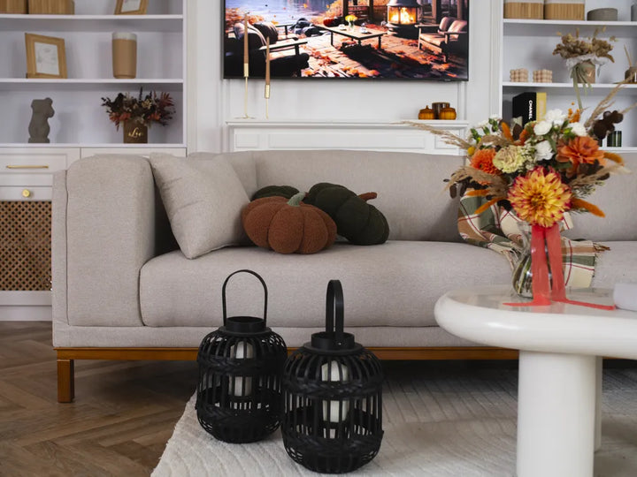 A styled living room scene featuring a light beige fabric sofa with decorative pumpkins, a white side table with a colorful floral arrangement, and two black lanterns in the foreground.