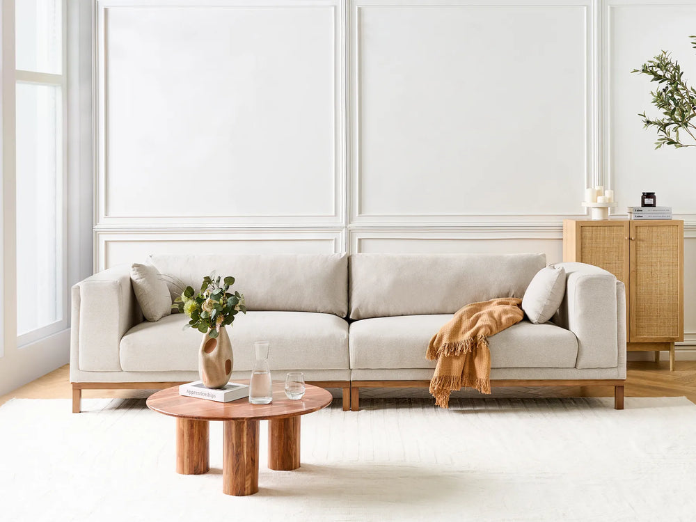 A light beige fabric sofa with a low wooden frame, styled in a bright, modern living room with a round wooden coffee table and a draped orange throw blanket.
