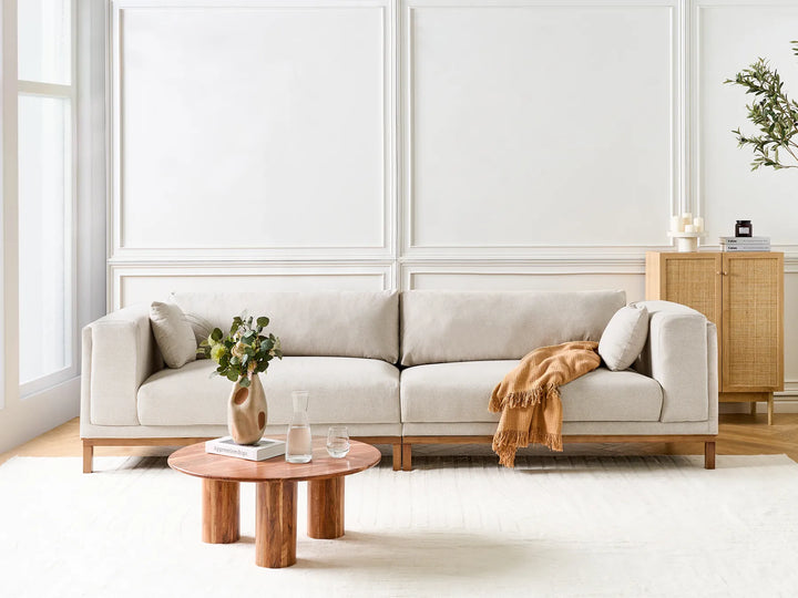 A light beige fabric sofa with a low wooden frame, styled in a bright, modern living room with a round wooden coffee table and a draped orange throw blanket.