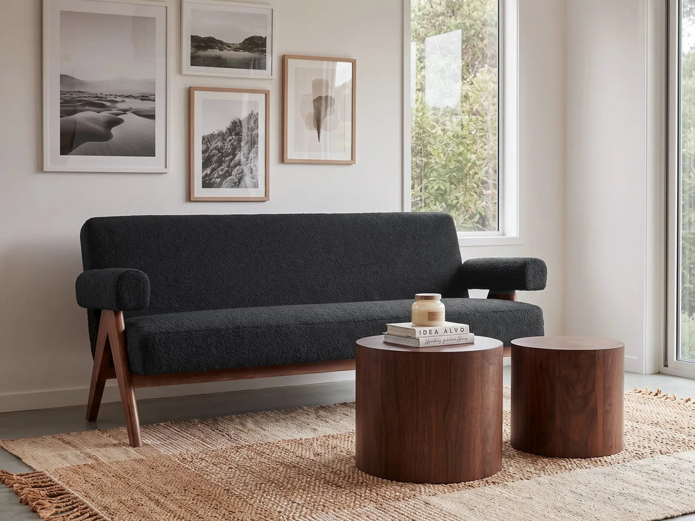 A dark charcoal gray upholstered sofa with angled wooden legs sits in a bright, modern living room setting with two matching wooden side tables, a woven rug, and framed wall art.