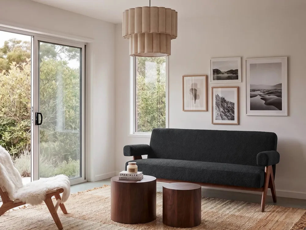 A modern living room scene featuring a dark charcoal gray upholstered sofa with wooden legs, two round dark wood side tables, a white sheepskin chair, and a beige pleated pendant light hanging above. The room has off-white walls, framed black and white landscape photographs, and large windows looking out onto greenery.