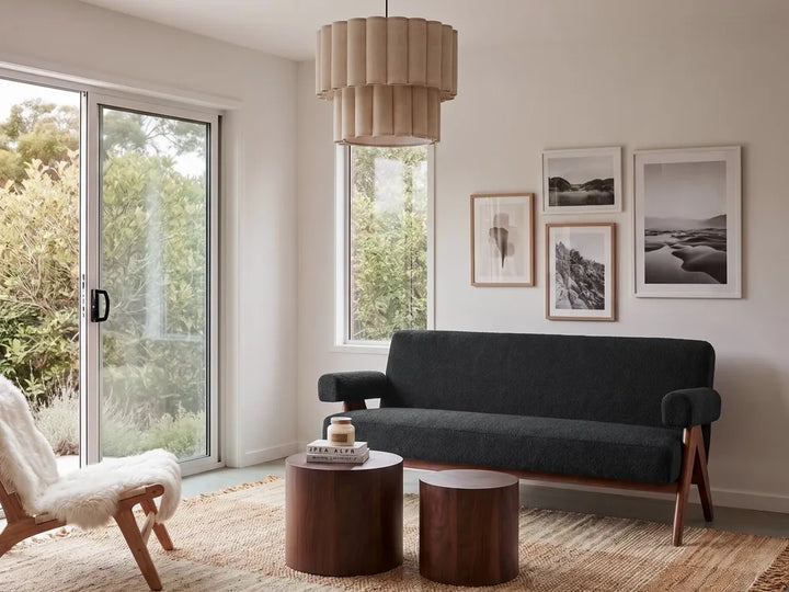 A modern living room scene featuring a dark charcoal gray upholstered sofa with wooden legs, two round dark wood side tables, a white sheepskin chair, and a beige pleated pendant light hanging above. The room has off-white walls, framed black and white landscape photographs, and large windows looking out onto greenery.