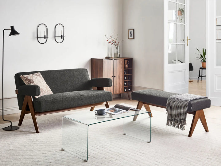 A mid-century modern two-seater sofa in dark gray fabric with angled wooden legs, shown in a bright, contemporary living room alongside a matching bench, glass coffee table, and wall sconces.