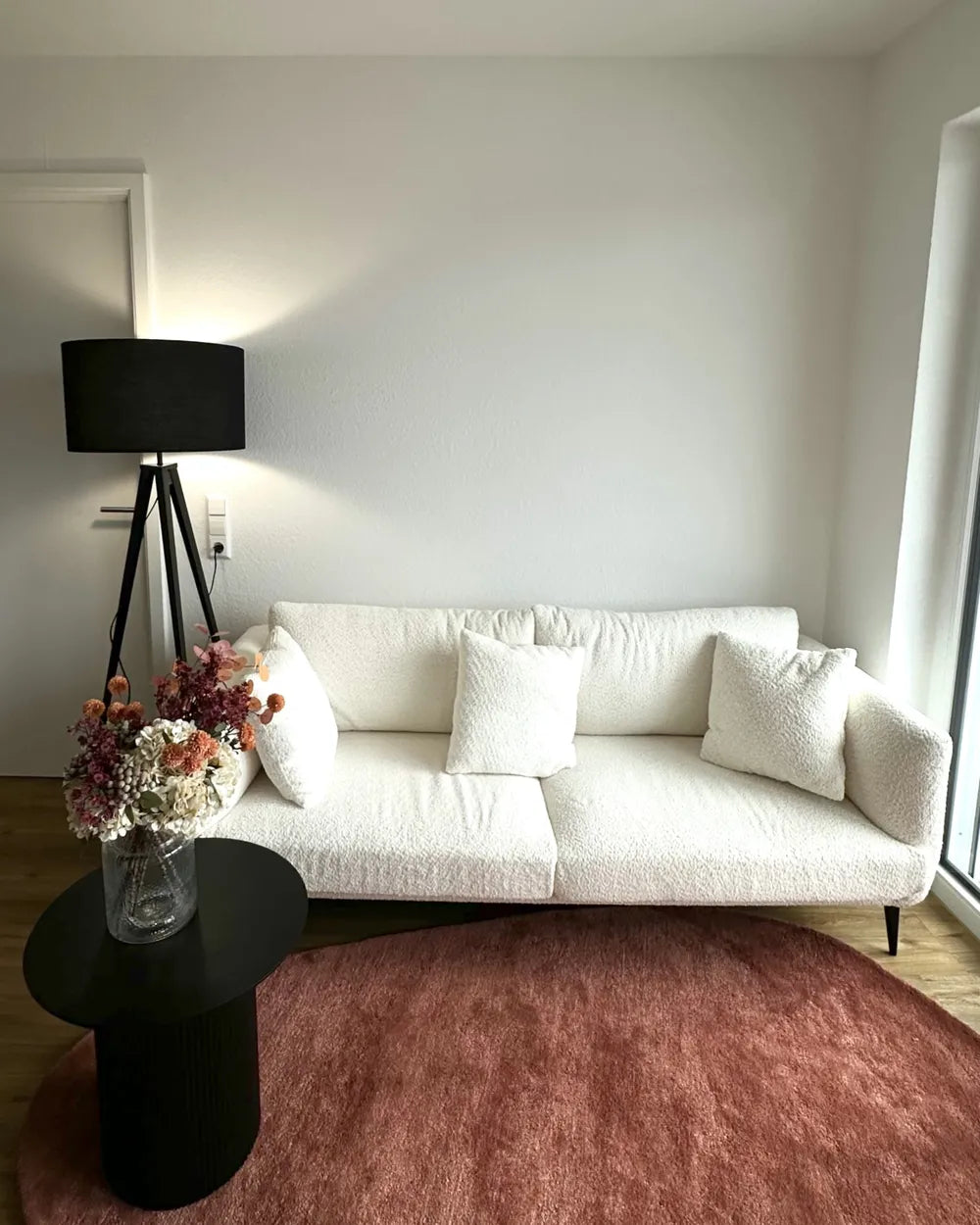 A modern living room scene featuring a white bouclé sofa with textured white throw pillows, a black tripod floor lamp, a small black side table with a vase of dried flowers, and a dusty rose round area rug on a light wood floor.