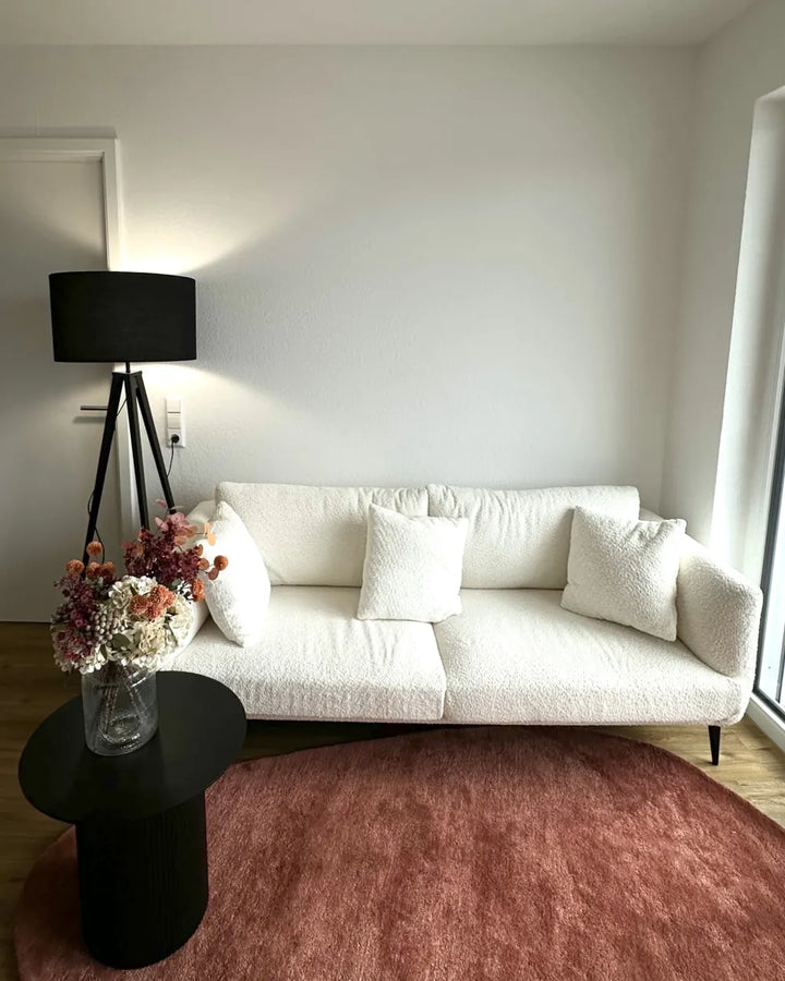 A modern living room scene featuring a white bouclé sofa with textured white throw pillows, a black tripod floor lamp, a small black side table with a vase of dried flowers, and a dusty rose round area rug on a light wood floor.