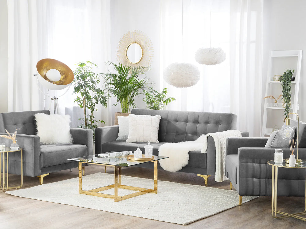 A modern living room setup featuring a gray tufted sofa, two matching armchairs, a glass-top coffee table with gold legs, a white rug, and decorative plants against a bright white curtain background.