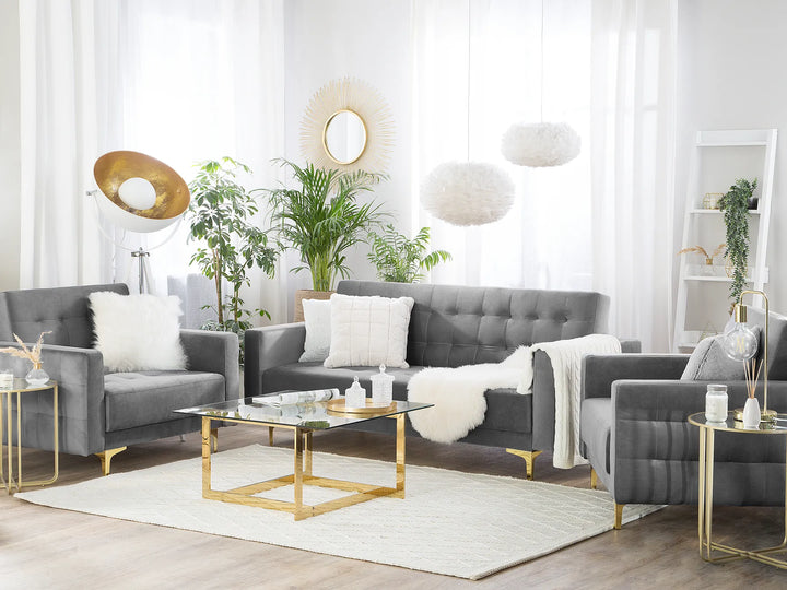 A modern living room setup featuring a gray tufted sofa, two matching armchairs, a glass-top coffee table with gold legs, a white rug, and decorative plants against a bright white curtain background.