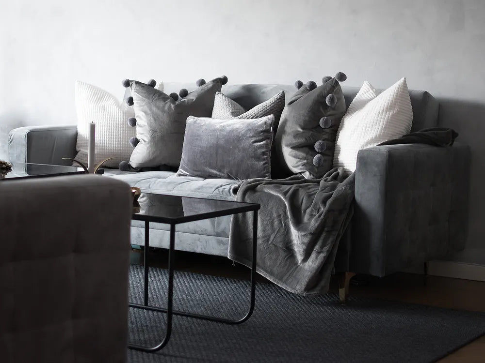 A gray velvet sofa in a modern living room setting, styled with multiple gray and white throw pillows, a matching gray throw blanket, and a black glass coffee table in the foreground.