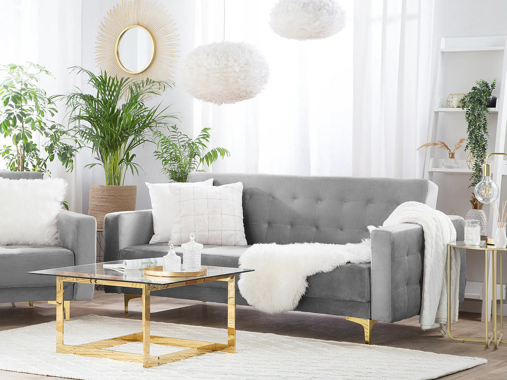 A modern gray tufted sofa in a bright, contemporary living room, styled with white throw pillows, a white faux-fur throw blanket, a glass-top coffee table with gold legs, and potted green plants.