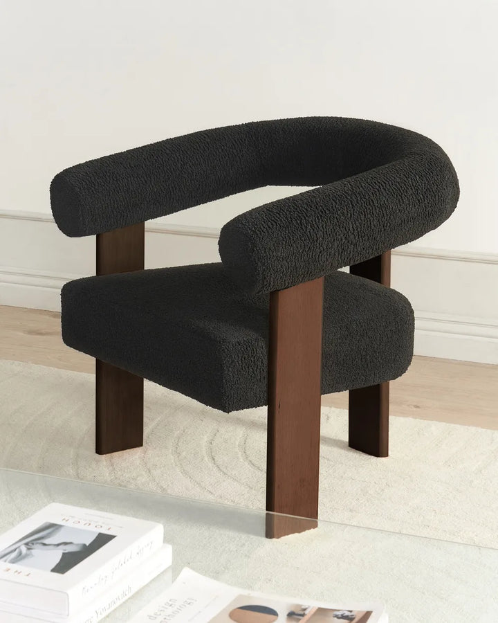 A modern accent chair with a dark charcoal bouclé upholstered seat and curved armrests, supported by dark brown wooden legs, shown in a minimalist room setting.