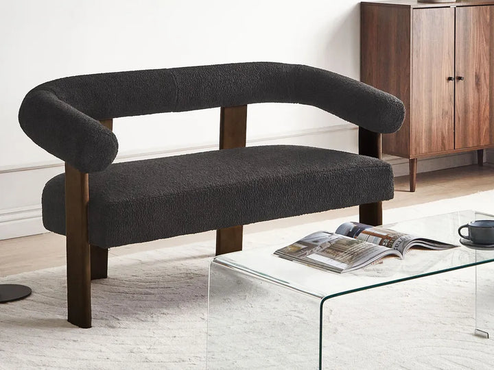 A modern curved bench with dark charcoal bouclé upholstery and dark wood legs, styled in a living room setting next to a glass coffee table.