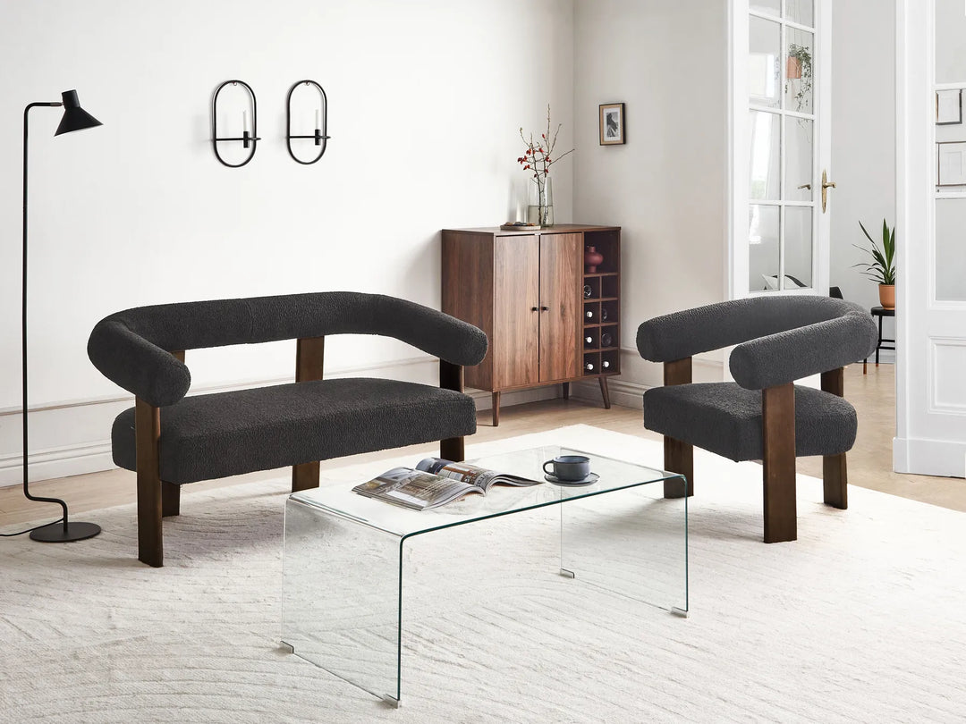 A modern living room furniture set featuring a dark gray upholstered loveseat, a matching armchair, a clear glass coffee table, and a wooden sideboard cabinet in a bright, minimalist room.