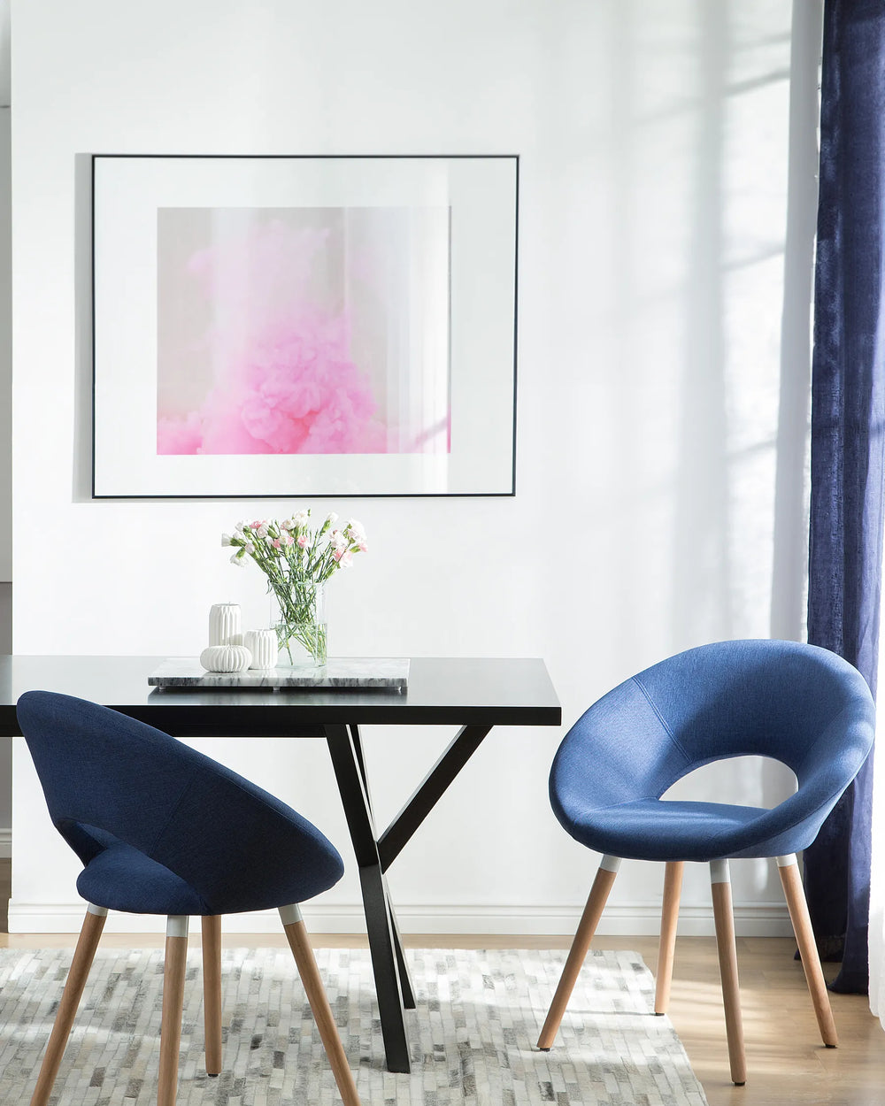 Modern interior with two blue chairs around a black table, framed artwork on the wall, and a vase with flowers.