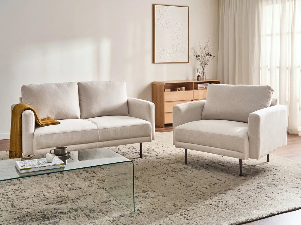 A modern living room setup featuring a light beige upholstered loveseat and matching armchair on a textured area rug, with a glass coffee table in the foreground and a wooden cabinet against the wall.