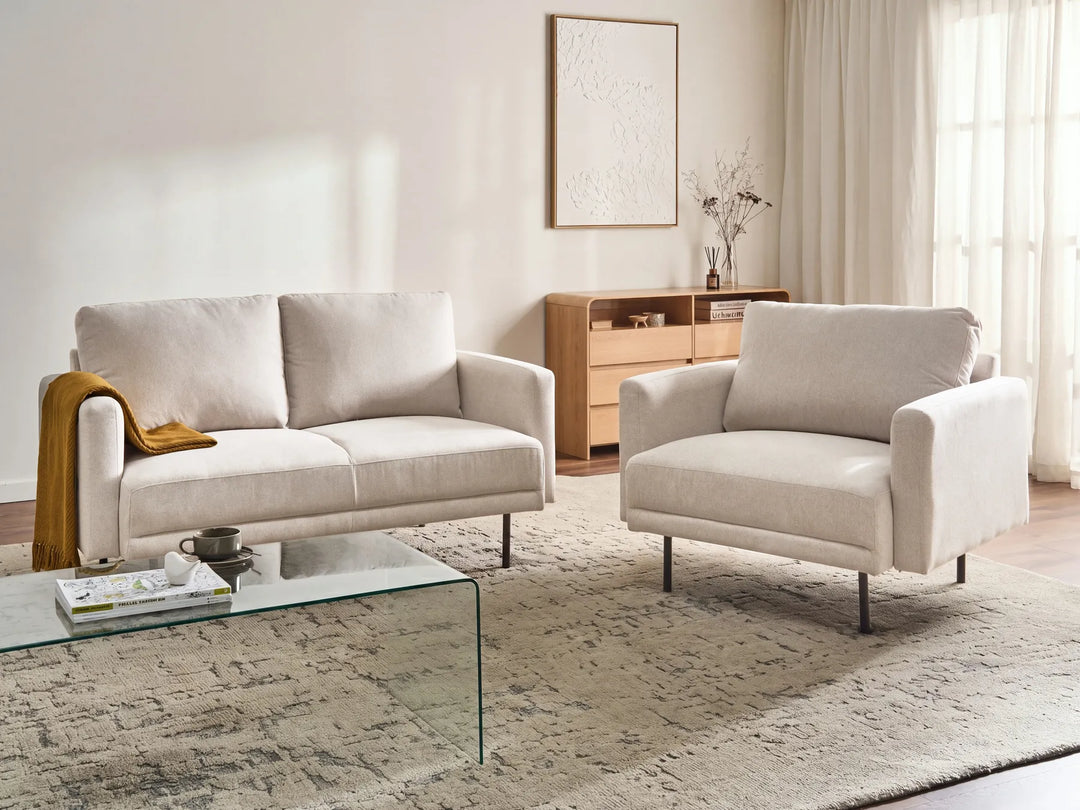 A modern living room setup featuring a light beige upholstered loveseat and matching armchair on a textured area rug, with a glass coffee table in the foreground and a wooden cabinet against the wall.