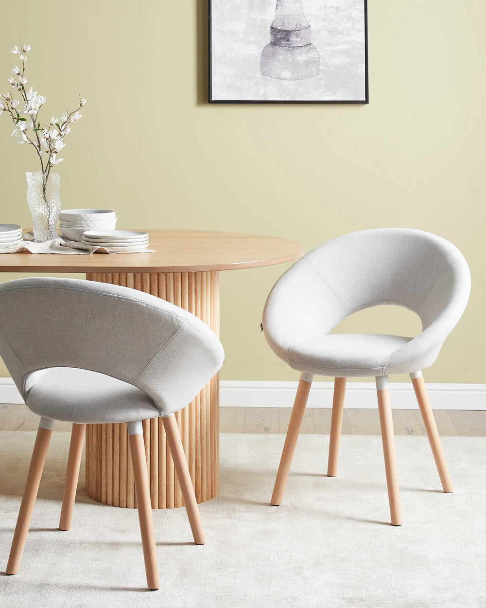 Two modern upholstered dining chairs with light gray fabric and natural wood legs, shown in a styled dining room setting next to a round wooden table.