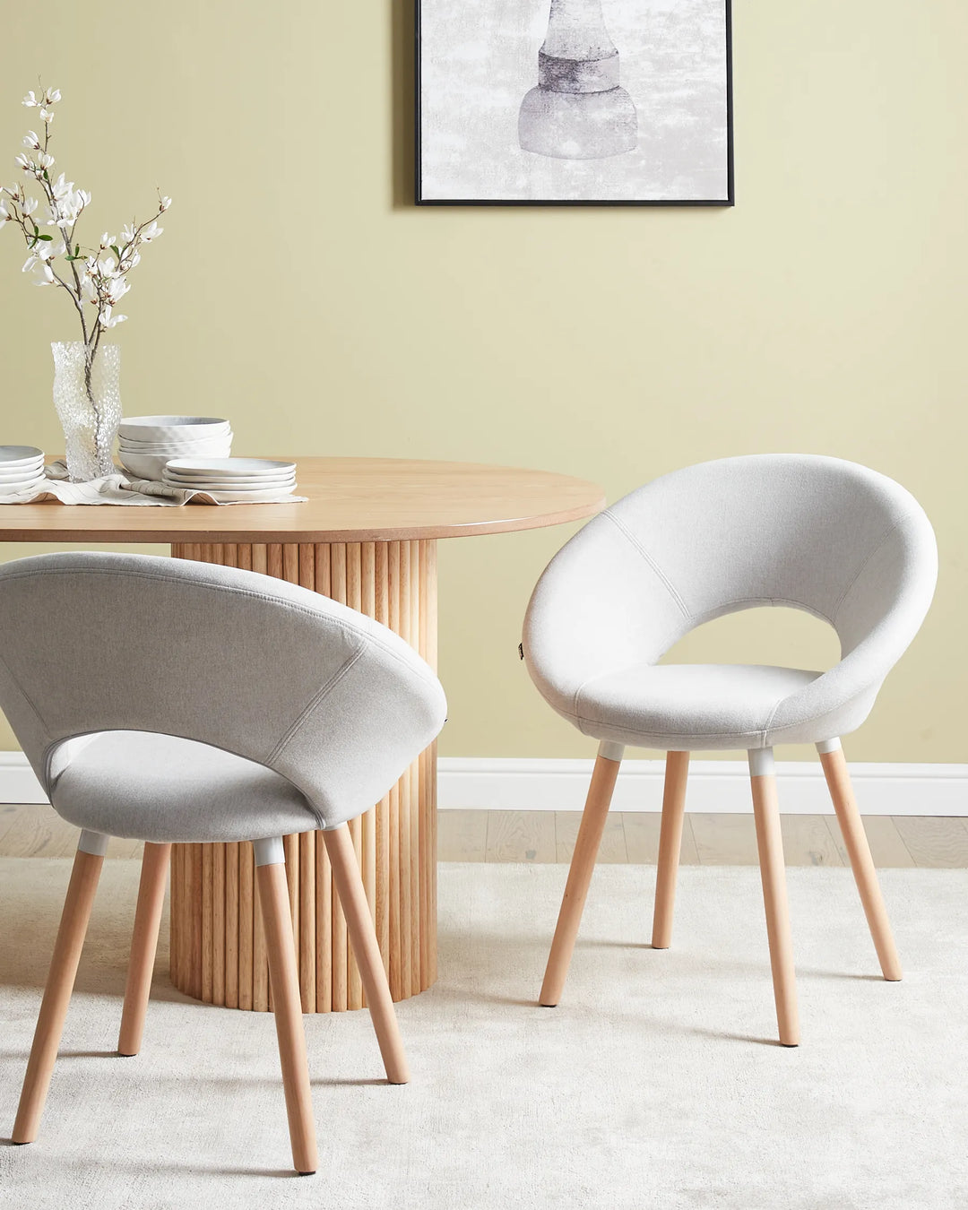 Two modern upholstered dining chairs with light gray fabric and natural wood legs, shown in a styled dining room setting next to a round wooden table.