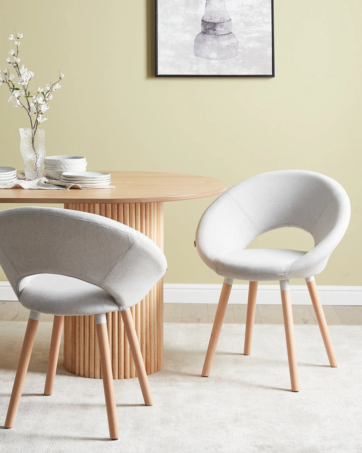 Two modern upholstered dining chairs with light gray fabric and natural wood legs, shown in a styled dining room setting next to a round wooden table.