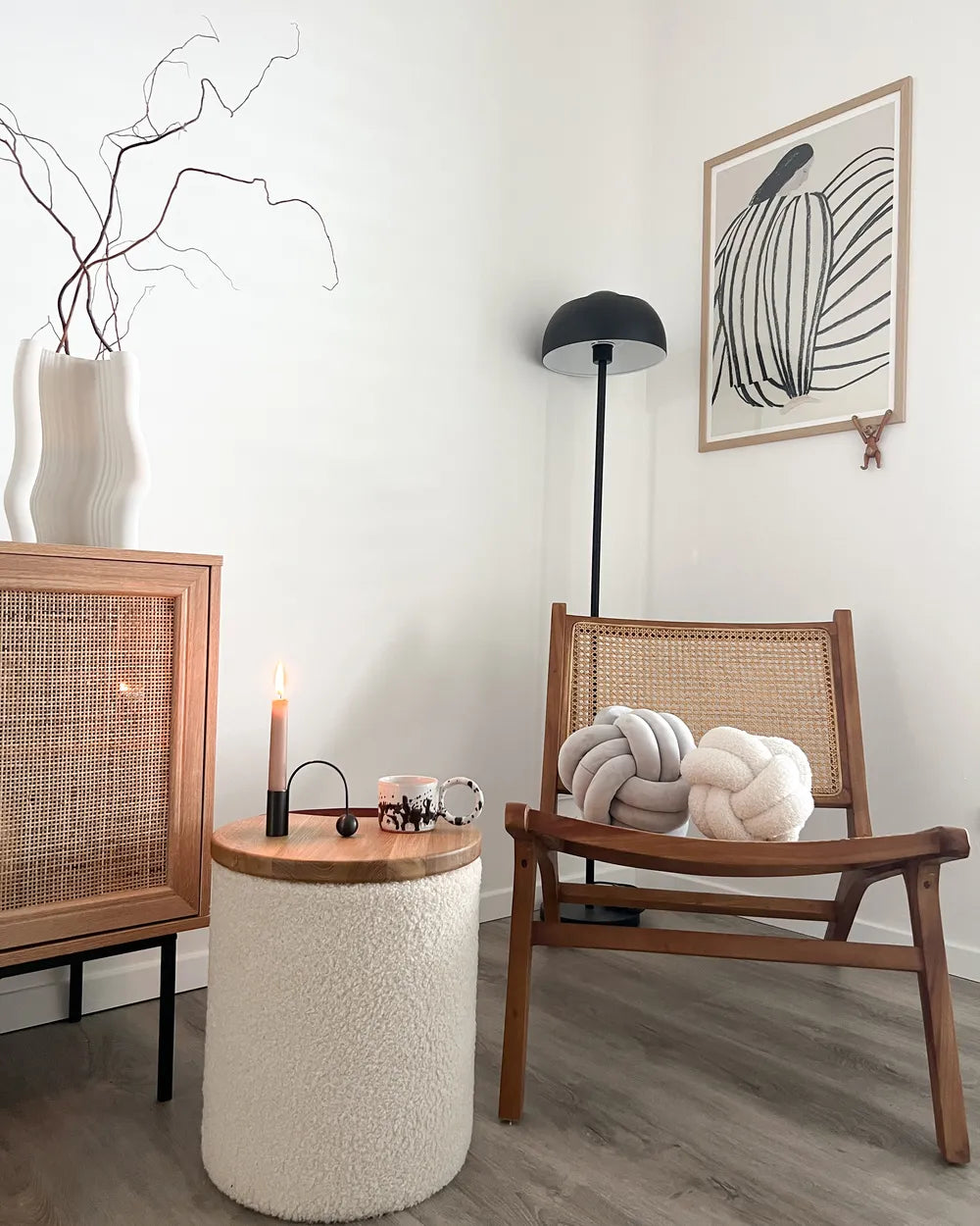 A cozy, modern living room corner featuring a mid-century wooden armchair with a woven cane backrest, a matching wooden sideboard with a cane front, a textured cream boucle side table with a lit candle, and a black floor lamp against a white wall.