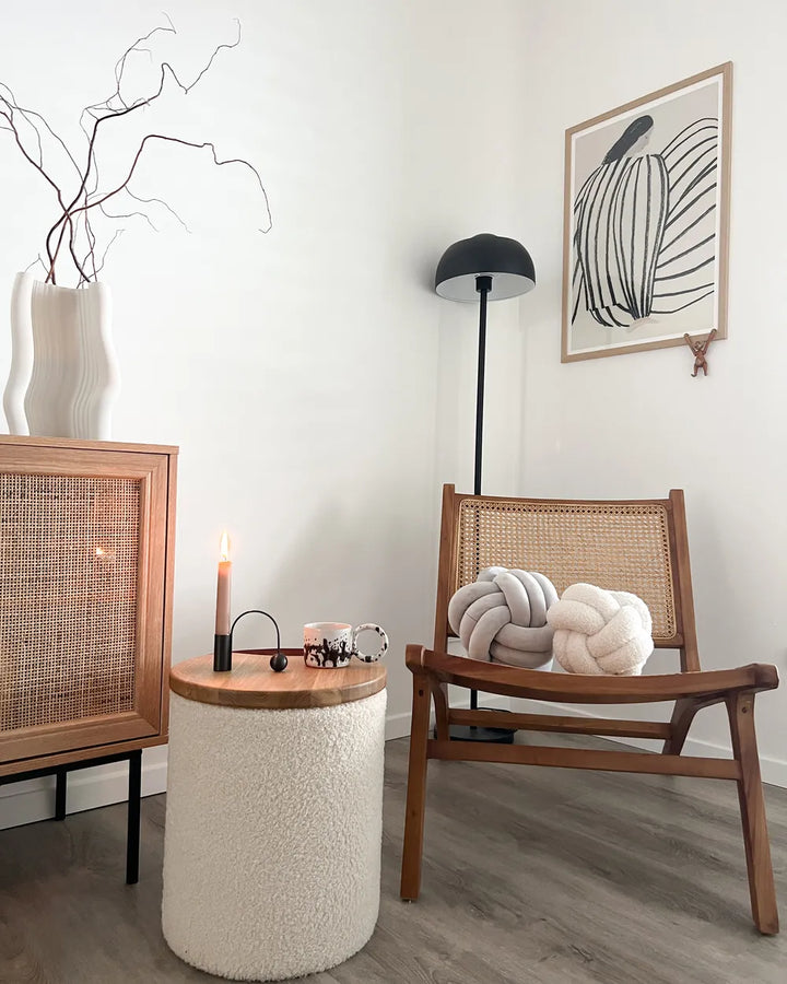 A cozy, modern living room corner featuring a mid-century wooden armchair with a woven cane backrest, a matching wooden sideboard with a cane front, a textured cream boucle side table with a lit candle, and a black floor lamp against a white wall.