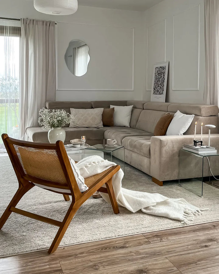 A styled living room featuring a large beige sectional sofa, a mid-century wooden cane armchair with a white throw blanket, a glass coffee table, and a light beige rug on a wood floor.